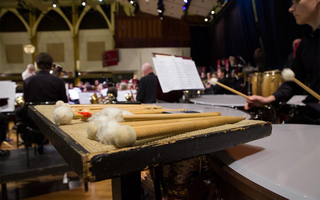 Timpani sticks on a traps table in front of a musician playing the timpanis. In the distance you can see an orchestra.